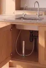 FINAL-1-vrysaki - Olympus Plus Open kitchen cabinet under the sink showing plumbing: pipes, a U-shaped trap, flexible hoses, and a small cylindrical water filter device mounted inside.