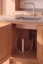 FINAL-2-triodi - Olympus Plus Open kitchen cabinet under the sink showing plumbing: a grayP-trap, white flexible hose, and vertical chrome pipe with a cylindrical device at the back.