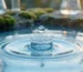 Macro shot of a water droplet creating a crown splash on a blue pool surface with ripples and blurred glassware in the background.